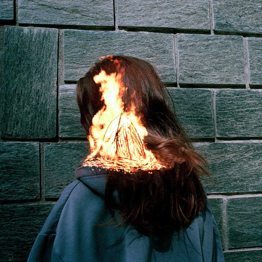 A young woman stands with her back toward us as she stands up close to a dark grey brick wall. Overlaid at head level against her hair are the flames of a burning wood fire.