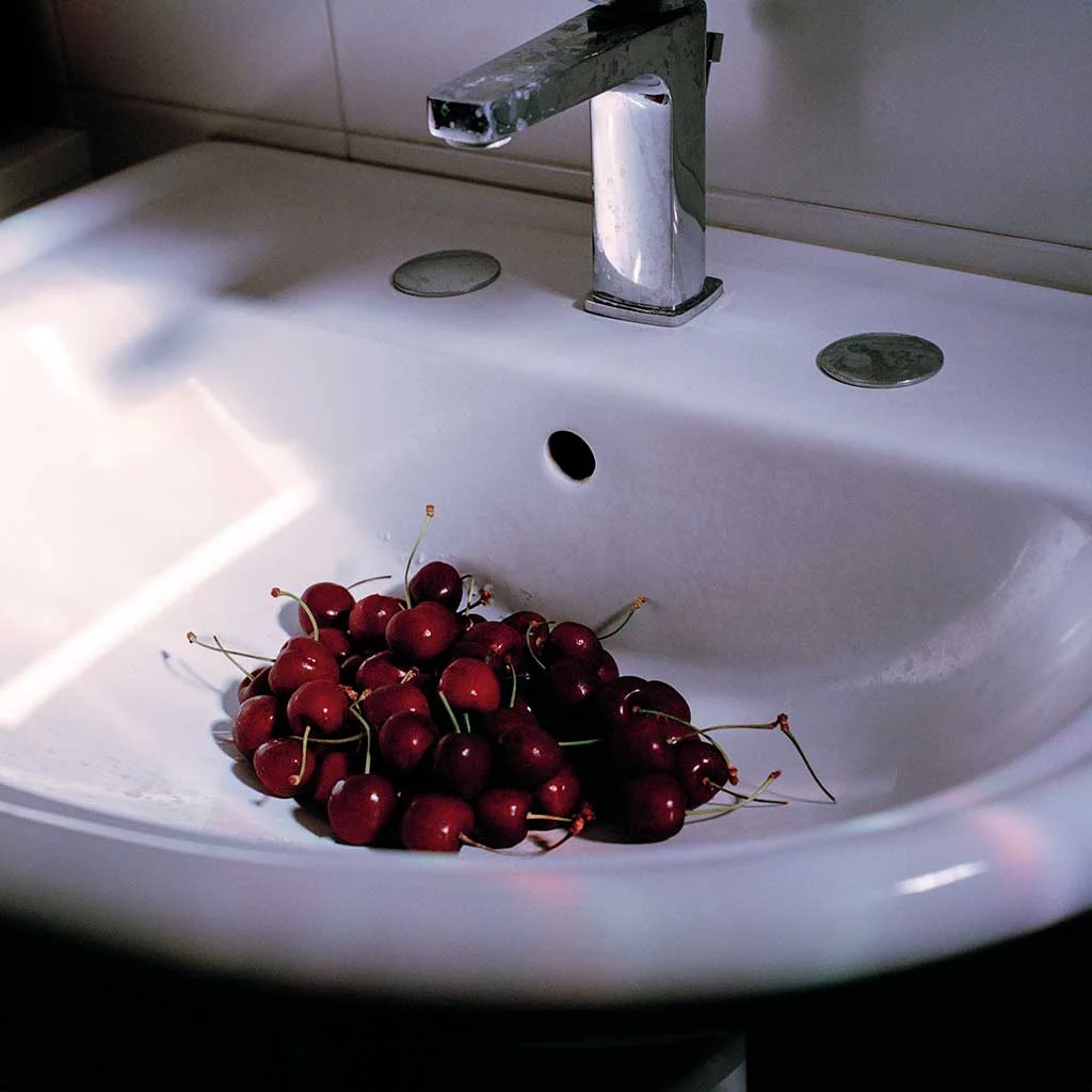 A pile of red cherries sits beneath the silver tap of a white bathroom washbasin. The fruit is in shadow while a beam of sunlight from an unseen window clips the edge of the white ceramic basin.