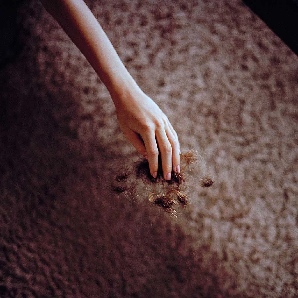 A young woman’s hand is dropping brown hair clippings that are falling toward a brown floor. The overall background is dark and blurred with a softly focussed light on the forearm and fingers.