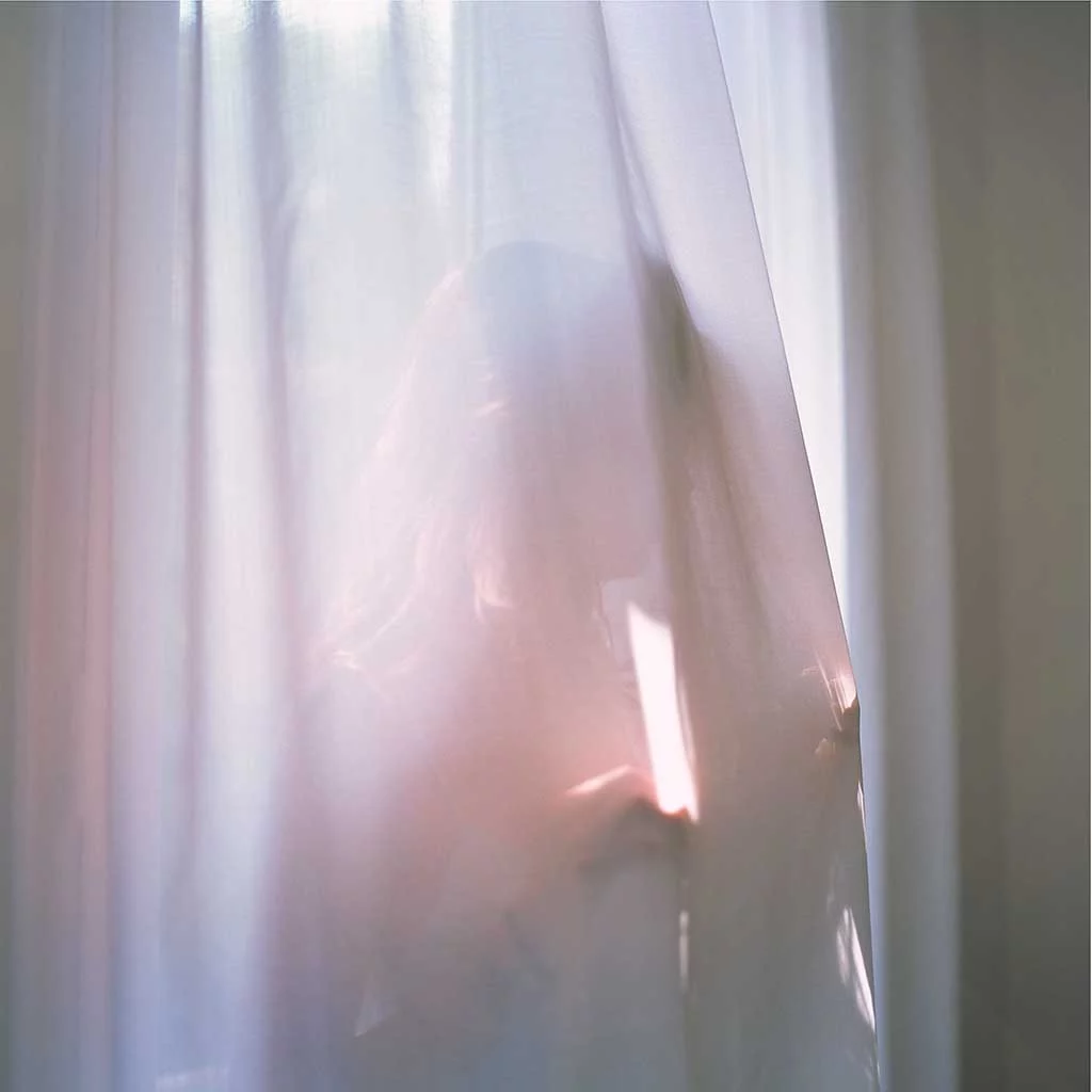 A girl in a sunny window standing behind a thin white curtain that is obscuring her face from view. Her hands lightly touch the fabric as she stand with her back to the window and looks through the curtain and in to the room.
