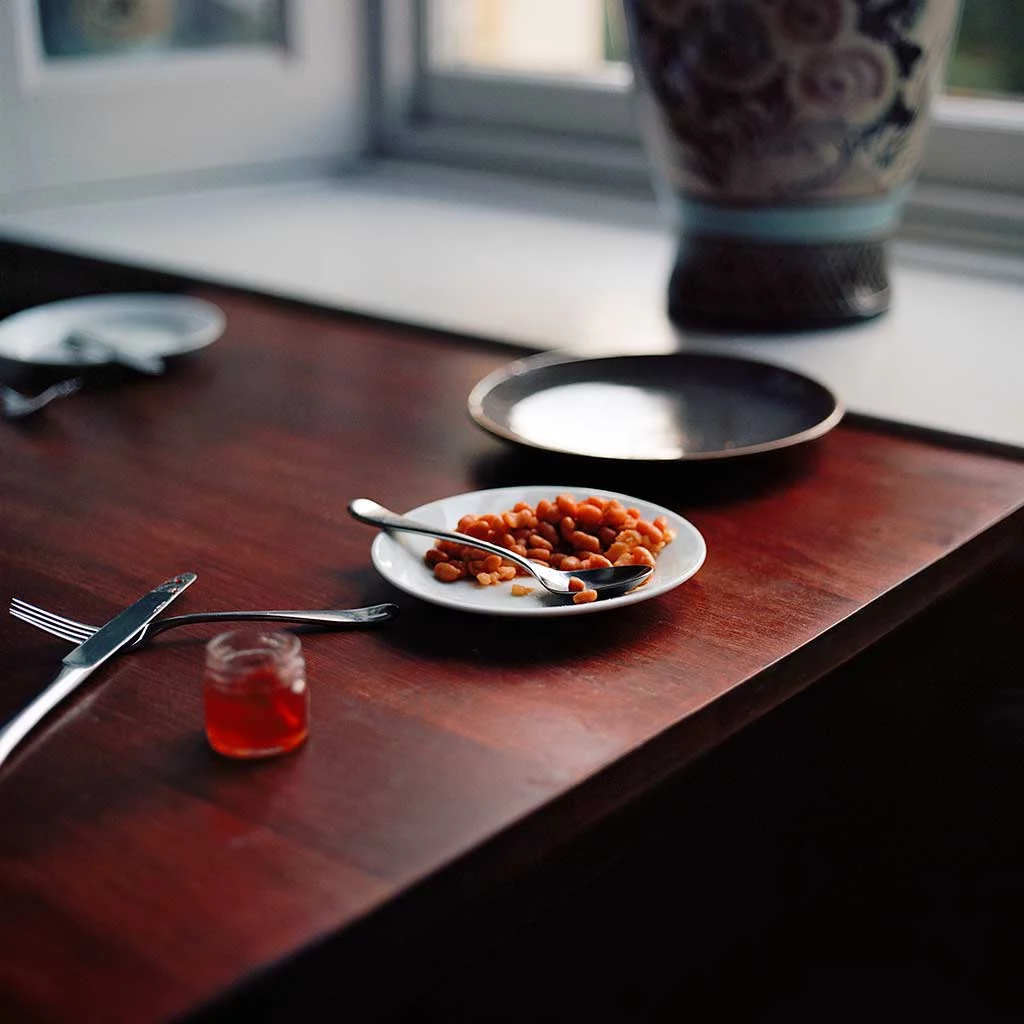 A table scene with discarded silver cutlery, small empty plates and food items. Set on a dark wooden table top.