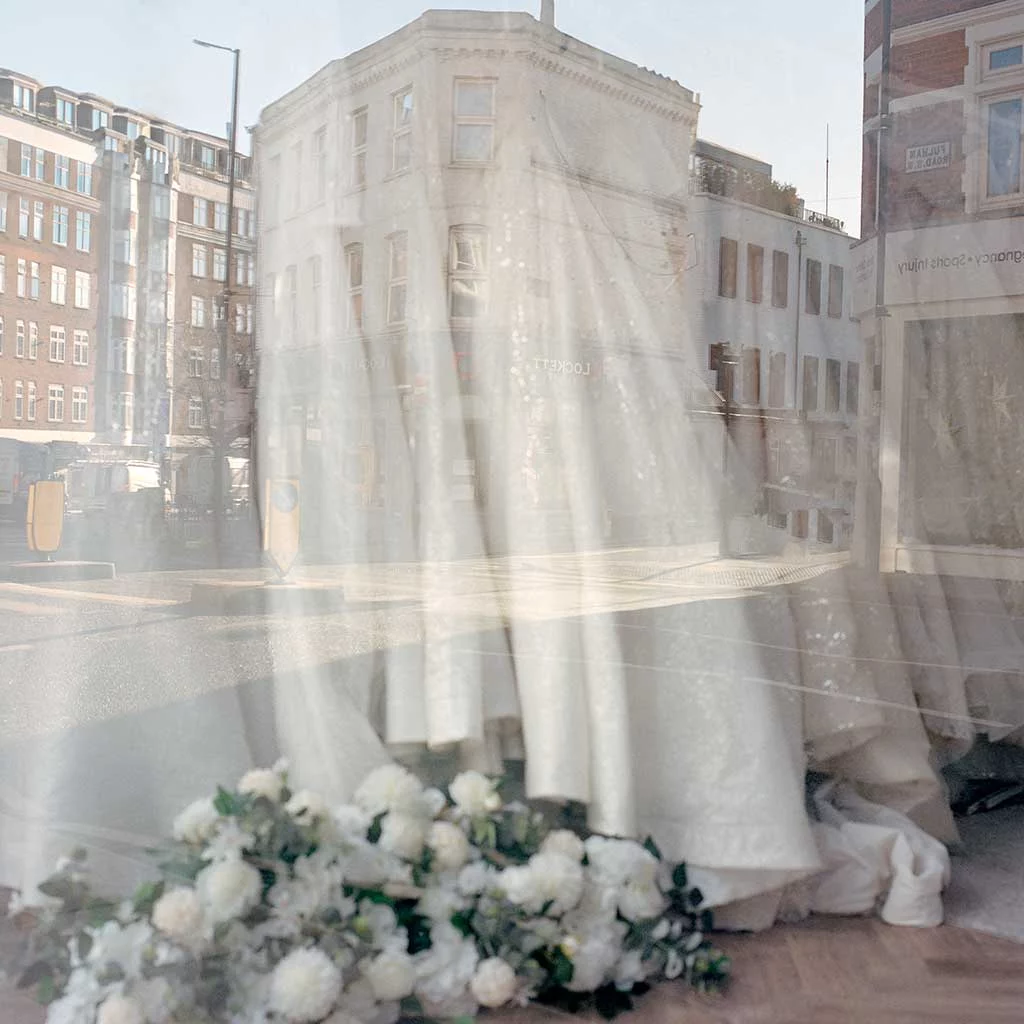 Reflections in the shop window of a bridal gown shop. White flowers and dresses hanging up inside the store blend and mix with the reflected city street scene.