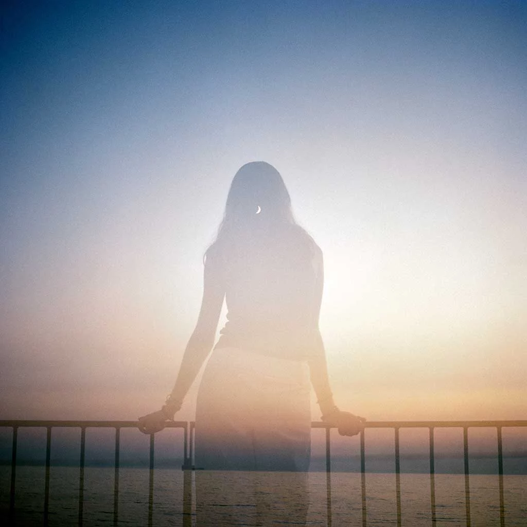 Reflected image showing the silhouette of a young woman leaning against metal railings and looking out to sea. Beyond her a crescent moon is visible the warm fading light of a cloudless evening sky.